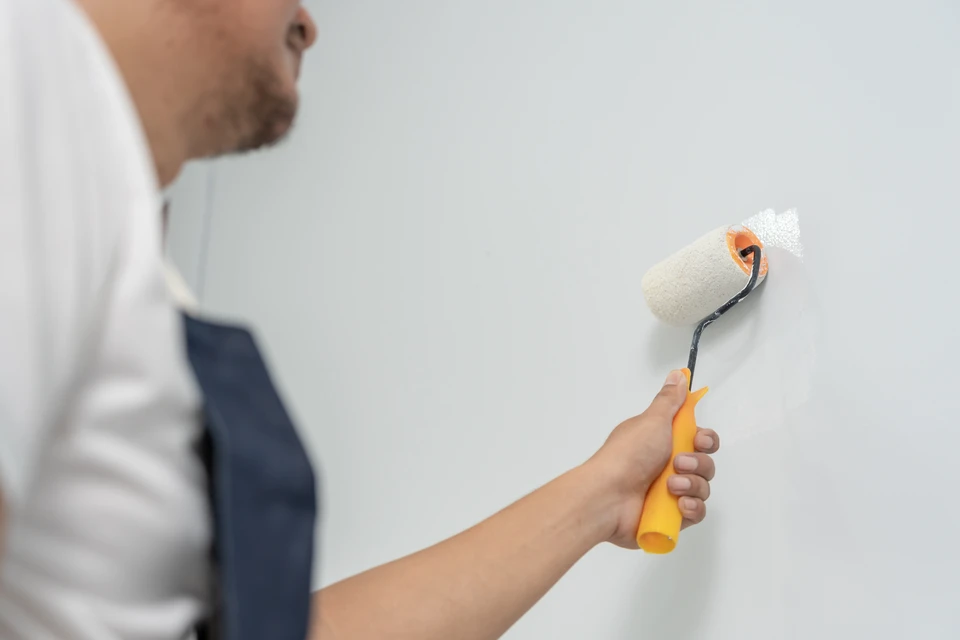 Artisan peintre en bâtiment portant tablier blanc et t-shirt bleu clair, tenant rouleau de peinture et faisant signe positif